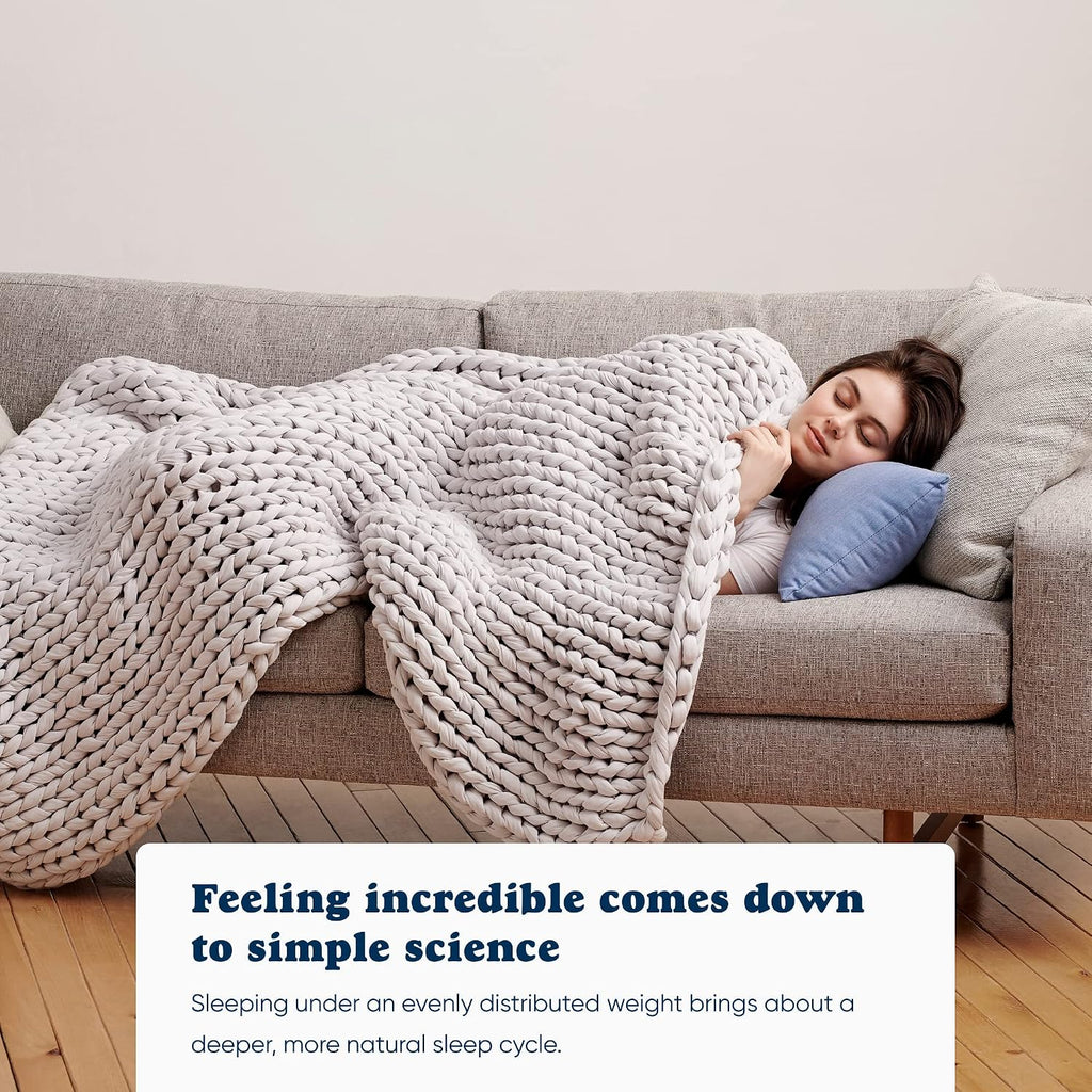 Woman napping on a grey couch under a light grey Bearaby Napper cotton weighted blanket, showing how evenly distributed weight supports deeper, more natural sleep