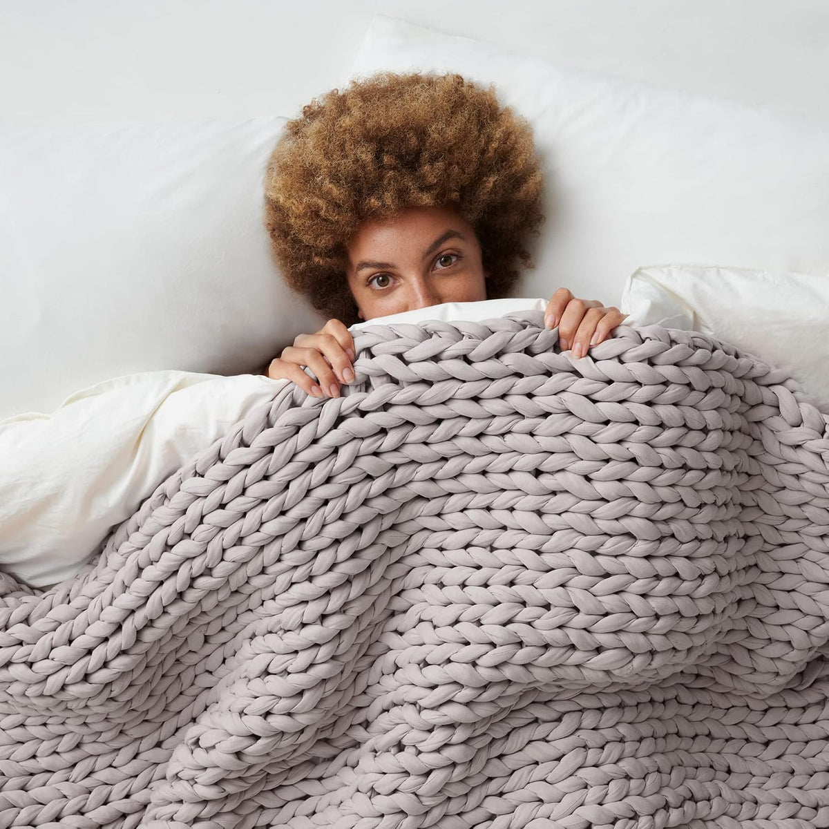 Woman in bed peeking over a light grey Bearaby Napper chunky knit cotton weighted blanket with white bedding