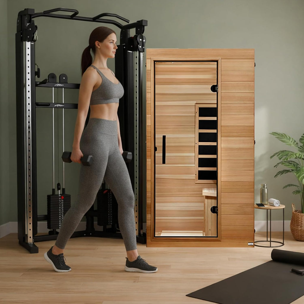 Dynamic Saunas Elite 1-person infrared cedar sauna cabin in a home gym beside a cable machine, woman walking past with dumbbells