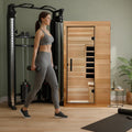 Dynamic Saunas Elite 1-person infrared cedar sauna cabin in a home gym beside a cable machine, woman walking past with dumbbells