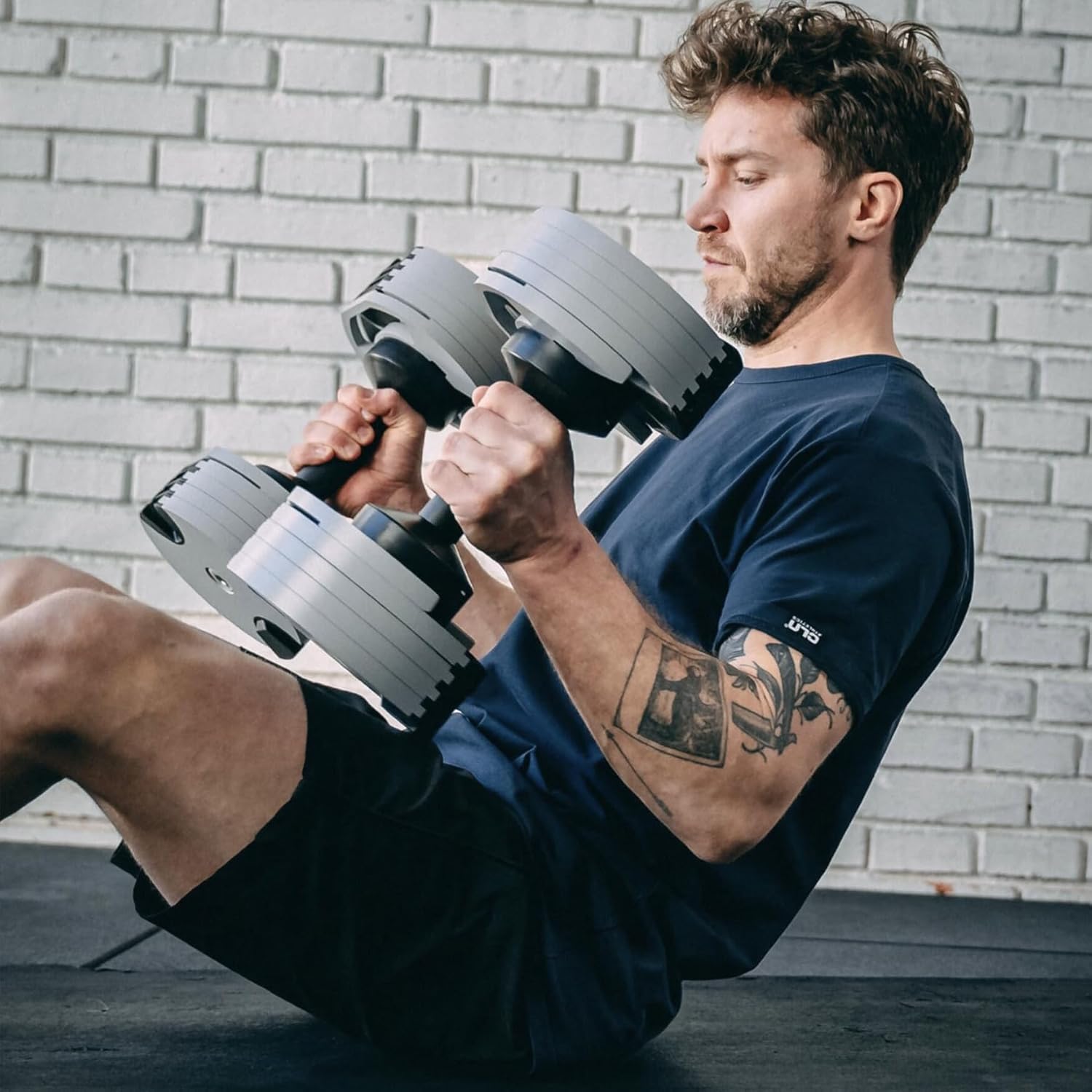 Man doing a seated ab exercise with NUOBELL adjustable dumbbells against a brick wall