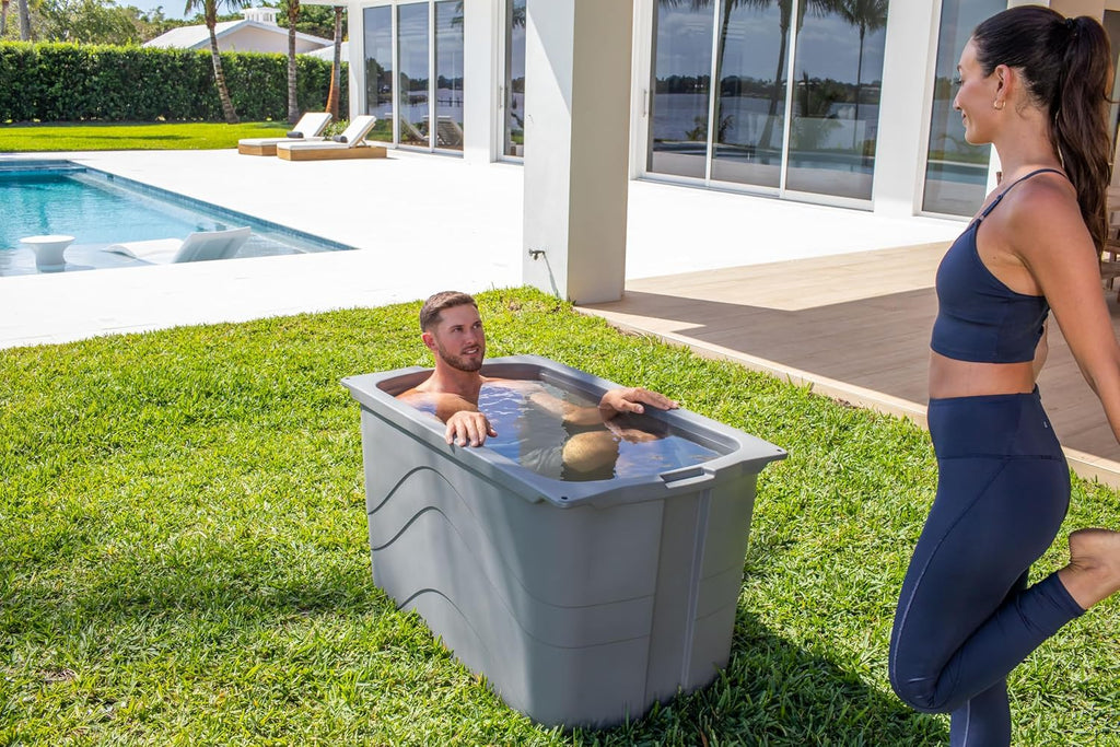 Man sitting in a grey Frozin 400 insulated cold plunge tub on a backyard lawn beside a pool, a woman stretching nearby