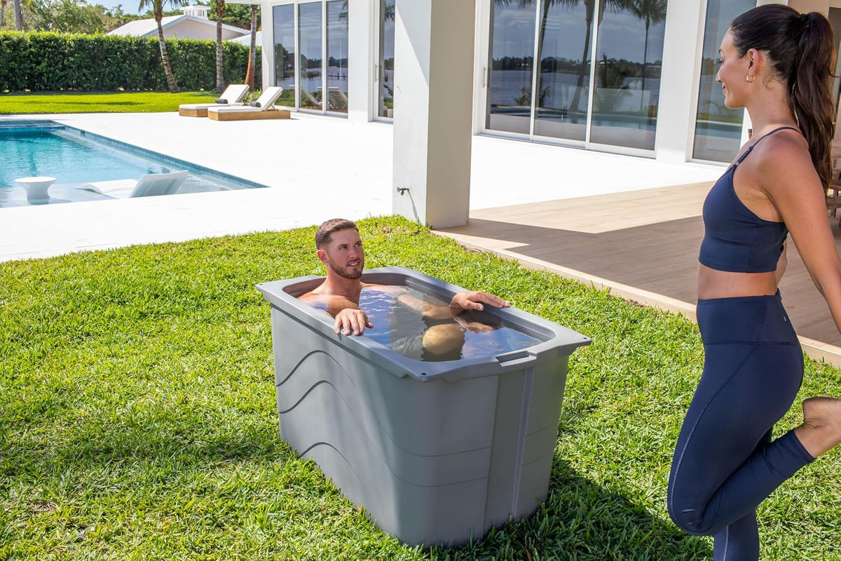 Man sitting in a grey Frozin 400 insulated cold plunge tub on a backyard lawn beside a pool, a woman stretching nearby