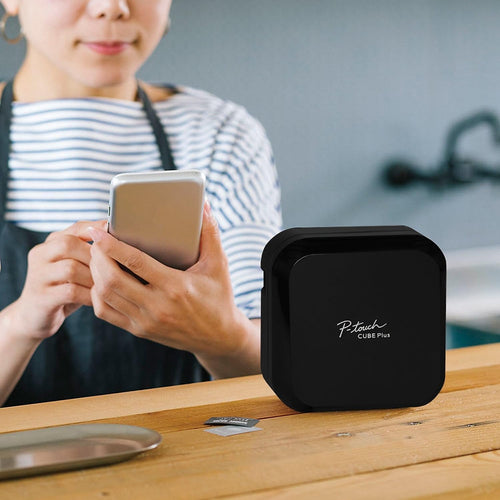 Woman using her phone to control the Brother P-touch CUBE Plus Label Maker on a wood table